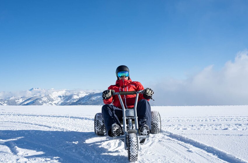 Person in red jacket riding snow kart on snow-covered terrain, mountains and blue sky in the background. | © Dorfgasteiner Bergbahnen AG