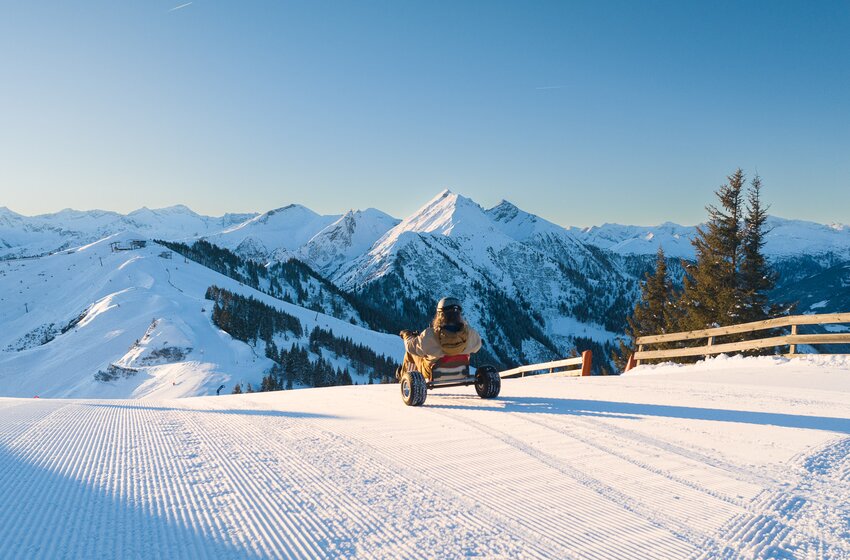 Person rides snow kart on freshly groomed slope with view of snowy mountains in bright sunshine. | © Dorfgasteiner Bergbahnen AG