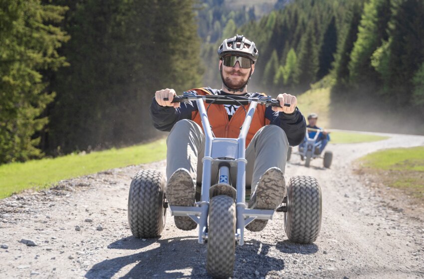 A man rides a mountain cart down a curving gravel path surrounded by forest and grass under bright sunshine | © Dorfgasteiner Bergbahnen AG