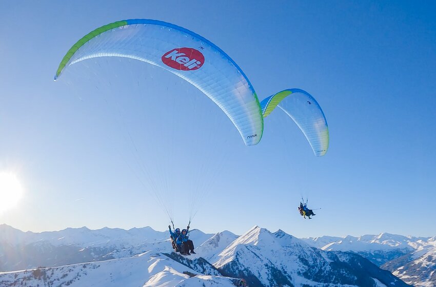Zwei Tandem-Paragleiter fliegen bei Sonnenschein über verschneite Alpenlandschaft mit blauem Himmel. | © Dorfgasteiner Bergbahnen AG