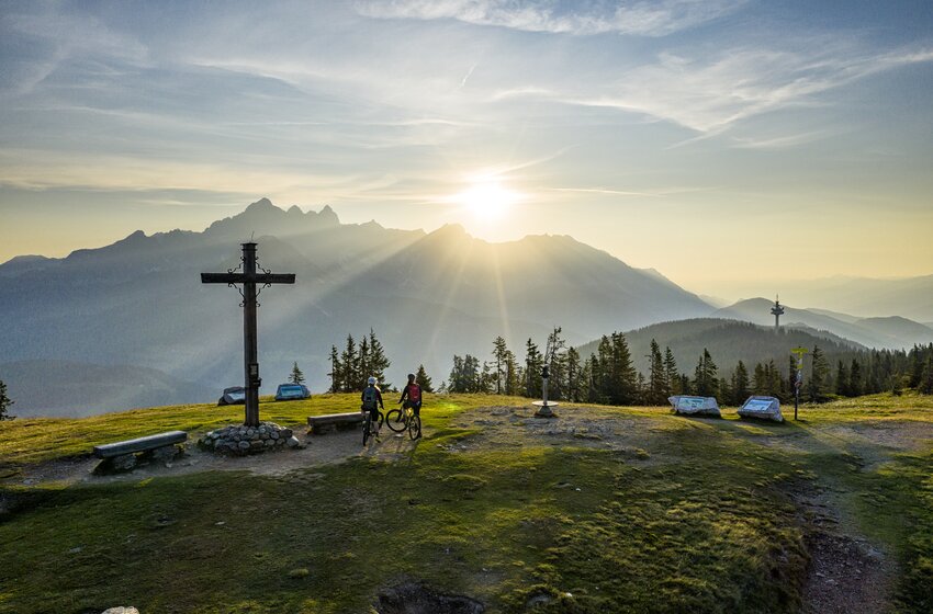 Zwei Mountainbiker stehen mit Rädern am Gipfelkreuz auf Wiese, Sonne über Bergkette, Wald und Fernsicht | © Bergbahnen Filzmoos GmbH