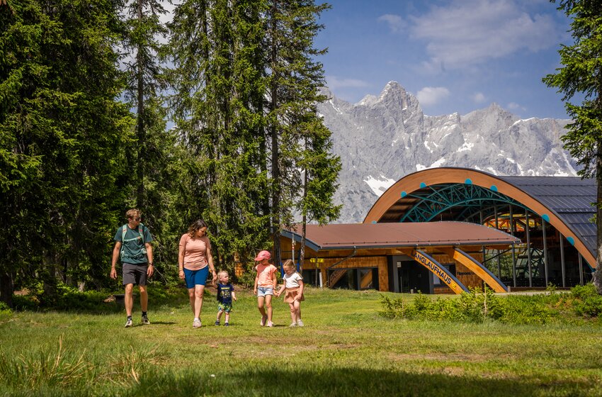 Zwei Erwachsene und drei Kinder gehen über Wiese vor Bergkulisse neben modernem Holzgebäude im Wald | © Bergbahnen Filzmoos GmbH