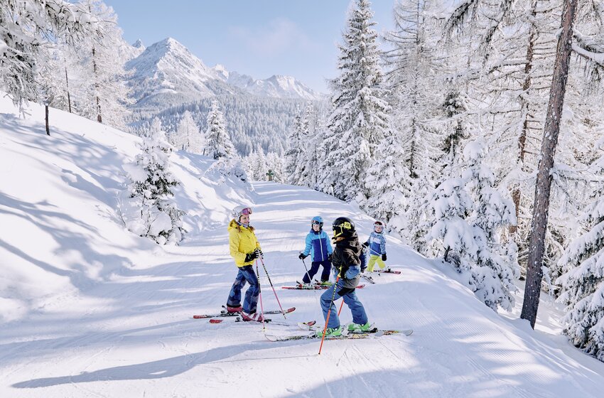 Four kids stand on a snowy forest slope, surrounded by trees and mountain scenery in winter. | © Bergbahnen Filzmoos GmbH