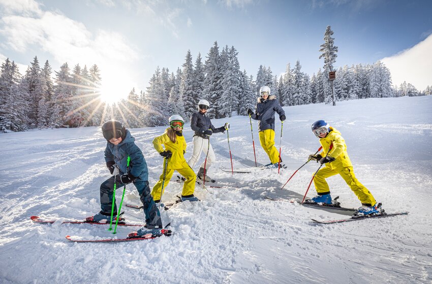 Fünf Skifahrer stehen mit Skiern auf einer verschneiten Piste vor winterlichem Nadelwald und tief stehender Sonne. | © Bergbahnen Filzmoos GmbH