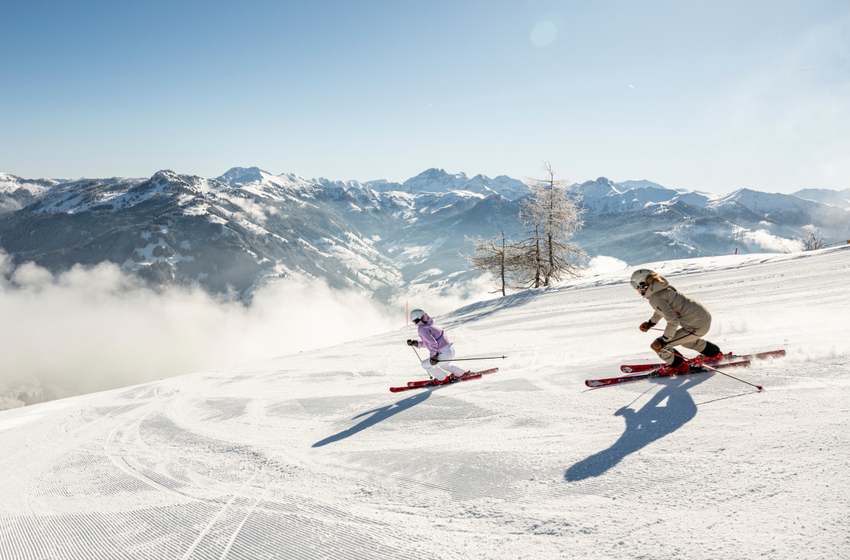 Two female skiers descend the sunny slope with mountain panorama in background | © Großarler Bergbahnen GmbH & Co KG