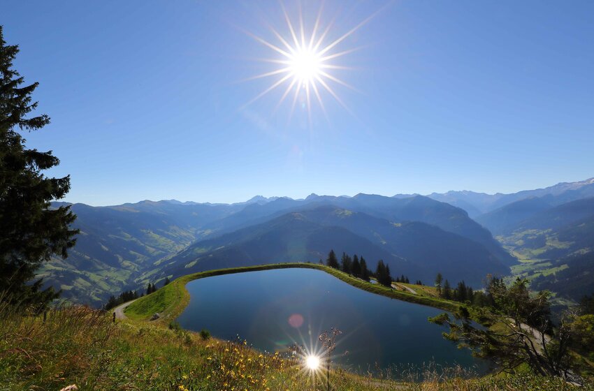 Reservoir lake in Großarltal with sun reflection and wide valley view | © Grossarler Bergbahnen GmbH & Co KG Mountain lake with sun reflection above Großarltal, overlooking green hills and distant alpine peaks | © Grossarler Bergbahnen GmbH & Co KG