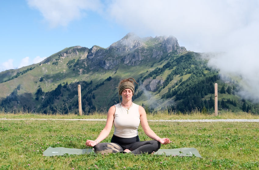 Eine Frau meditiert im Schneidersitz auf einer Yogamatte auf einer Wiese im Großarltal vor grünen Bergen. | © Grossarler Bergbahnen GmbH & Co KG