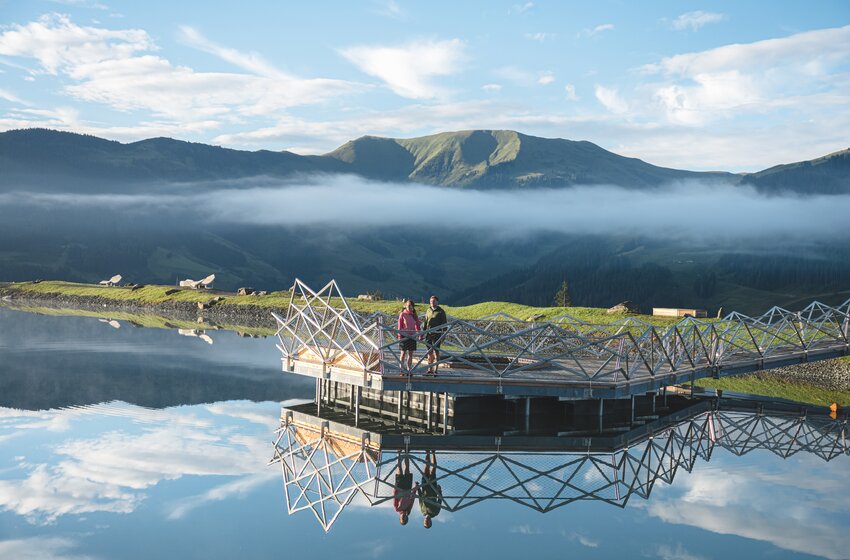Zwei Personen stehen auf Steg am ruhigen Bergsee mit Spiegelung vor Bergen und tiefen Wolken | © Hochkönig Bergbahnen GmbH