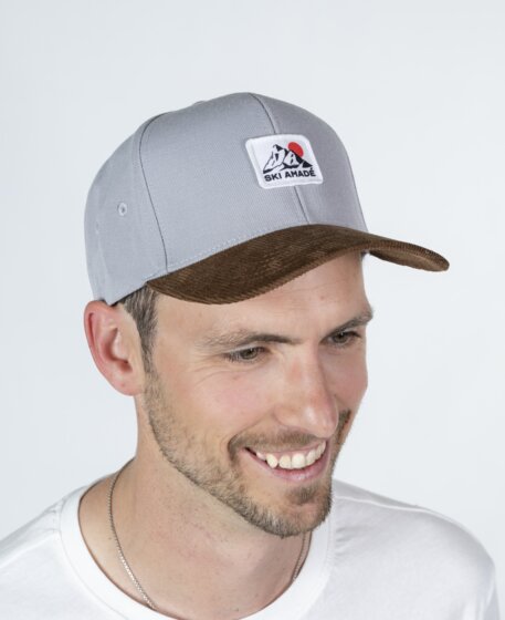 Man wearing a grey baseball cap with logo and brown brim and a white T-shirt in front of a light studio background | © Ski amadé GmbH / Lorenz Masser