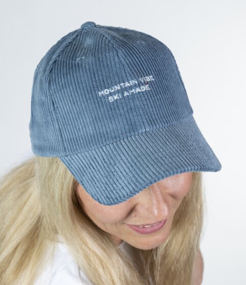 Blonde woman wearing a blue cord baseball cap with embroidery looking down in front of a light studio background | © Ski amadé GmbH / Lorenz Masser