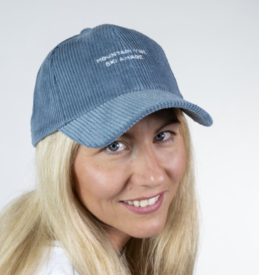Blonde woman wearing a blue cord baseball cap and white shirt smiling sideways in front of a light studio background | © Ski amadé GmbH / Lorenz Masser