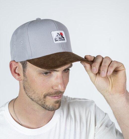 Man wearing grey baseball cap with Ski amadé logo and brown brim holding the cap in front of a light studio background | © Ski amadé GmbH / Lorenz Masser
