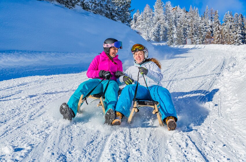 Two smiling women toboggan side by side down a snowy slope with snow spraying up around them. | © René Eduard Perhab