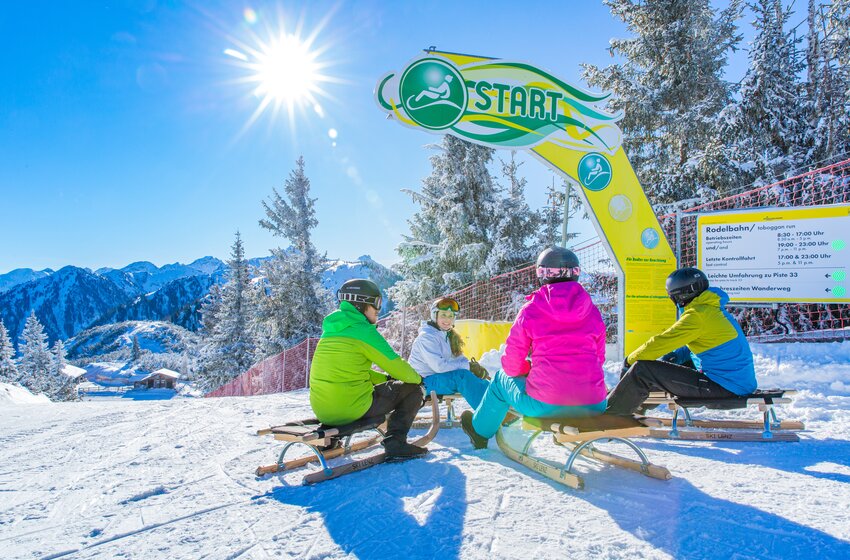 Four tobogganers sit at a sunny toboggan run start line with snowy mountain views in the background. | © René Eduard Perhab