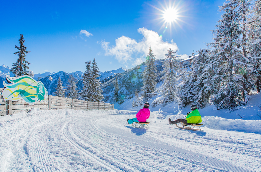 Two tobogganers ride through a sunny snowy curve with mountain and forest backdrop in brilliant winter light. | © René Eduard Perhab