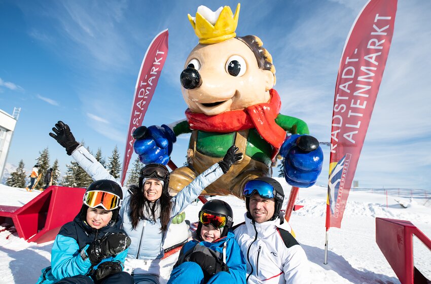 Family in ski gear posing cheerfully in front of mascot Kinigl in Radstadt-Altenmarkt ski area. | © Zauchensee Liftgesellschaft Benedikt Scheffer GmbH