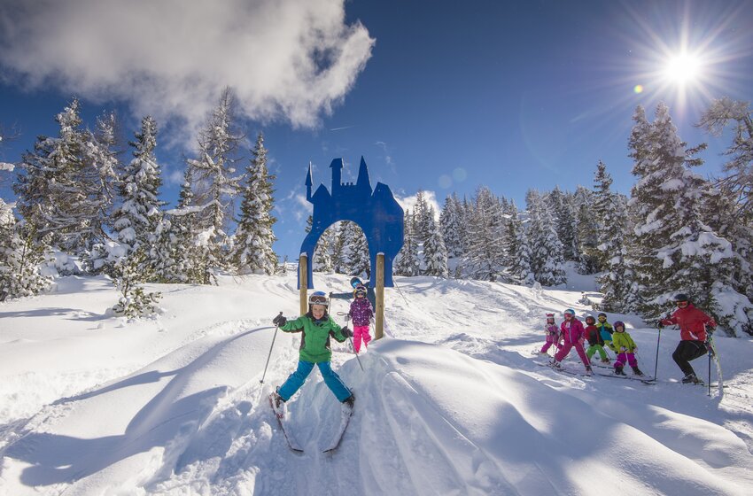 Kids skiing through a blue fairytale gate in the snow under bright sun and deep blue sky. | © Reiteralm Bergbahnen GmbH & Co.KG