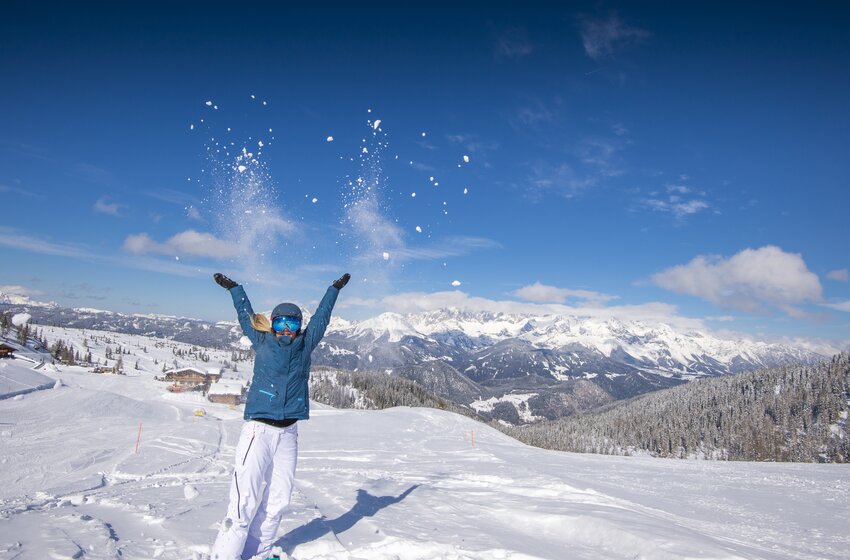 Woman throws snow into the air with snowy mountains and a bright blue sky behind her. | © Reiteralm Bergbahnen GmbH & Co.KG