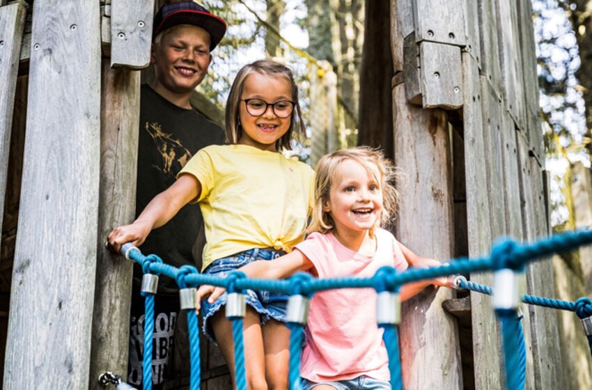 Kids’ forest adventure on Flori’s Nature Trail Griessenkareck | © Snow Space Salzburg Bergbahnen Two girls happily cross a rope bridge in the forest, with a smiling teenager standing behind them at a wooden play tower. | © Snow Space Salzburg Bergbahnen