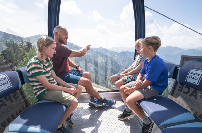 Family ride with the Flying Mozart gondola in Snow Space Salzburg | © Snow Space Salzburg Bergbahnen An adult sits with three children in the Flying Mozart gondola, looking out at the sunny mountain scenery outside. | © Snow Space Salzburg Bergbahnen