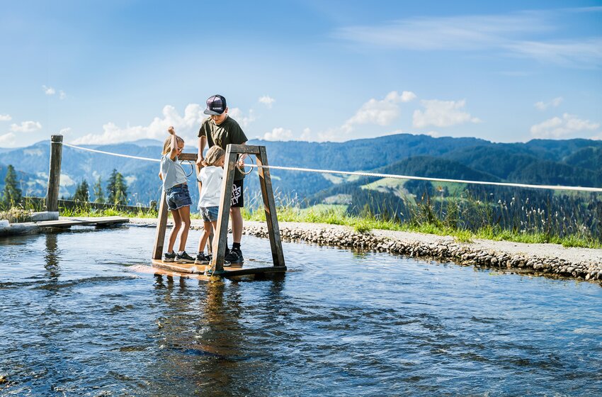 Three children stand on a water raft in a shallow pool with green hills and a clear summer sky behind. | © Snow Space Salzburg