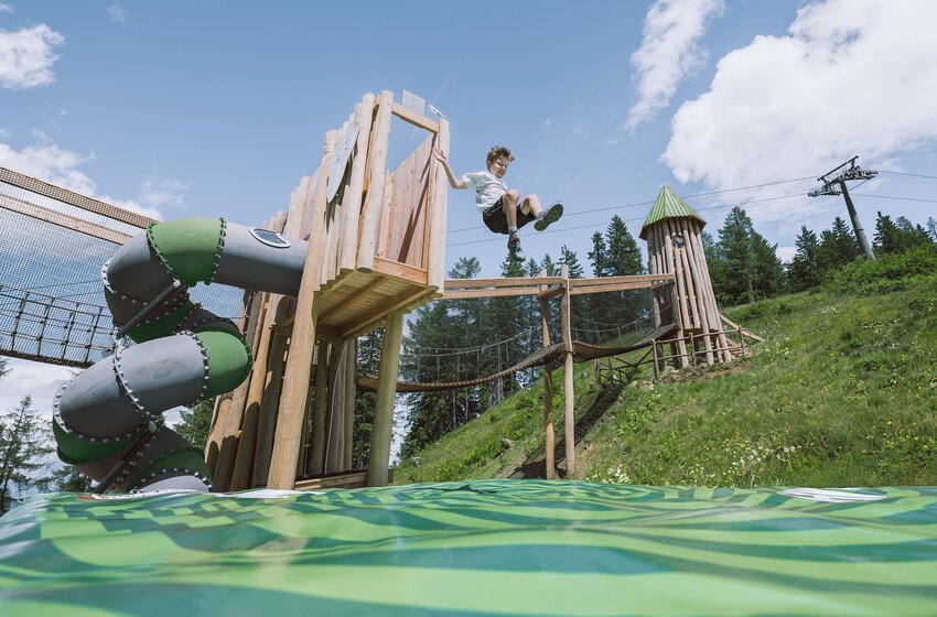 A child jumps from a wooden tower onto a jumping cushion beside a slide and climbing structures under a blue sky. | © Snow Space Salzburg