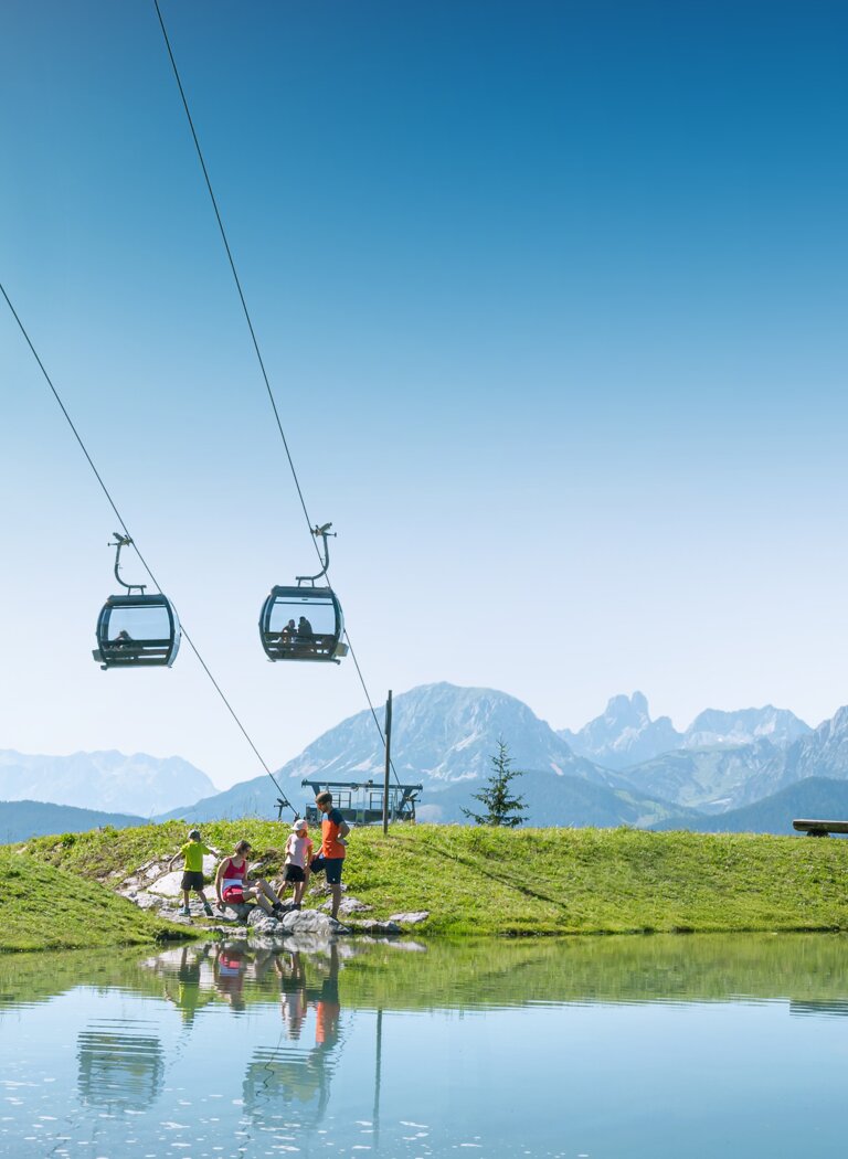 Family by the shore of a small mountain pond beneath a lift, with green meadows and Alps behind