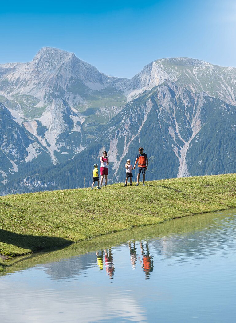 Family hiking by a clear mountain lake, with stunning peaks and reflections in the calm water. | © Ski amadé GmbH