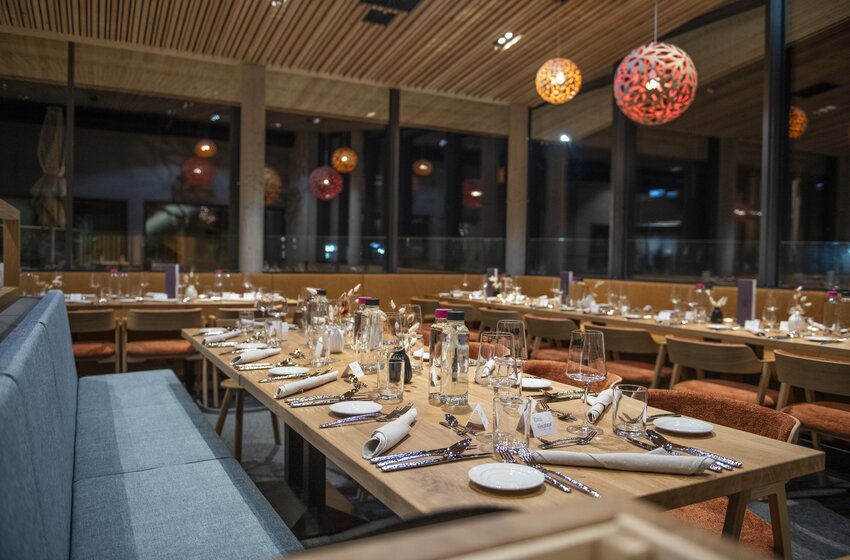 Modern restaurant with long set wooden tables, glasses, cutlery, bench seating and pendant lights beside large windows | © Gasteiner Bergbahnen AG 