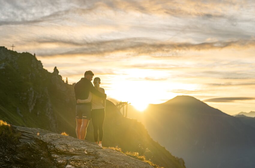 Paar steht eng umschlungen auf einem Felsen am Berggipfel, im Hintergrund Berge und leuchtender Himmel | © Gasteiner Bergbahnen AG