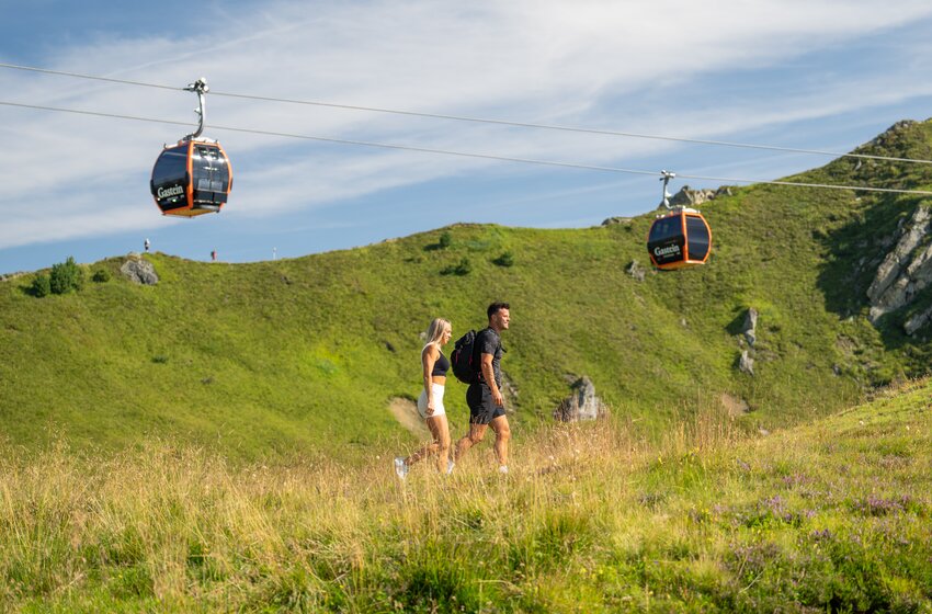 Couple hikes across green alpine meadow with backpack, cable cars above, surrounded by hills and blue sky | © Gasteiner Bergbahnen AG