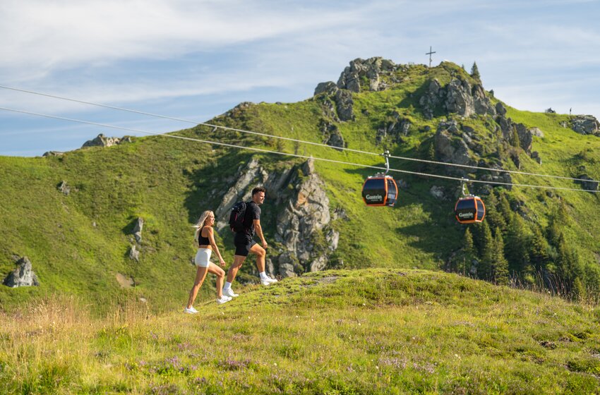 Paar wandert über grüne Almwiese mit Rucksack, im Hintergrund Gondeln und felsiger Gipfel mit Kreuz | © Gasteiner Bergbahnen AG