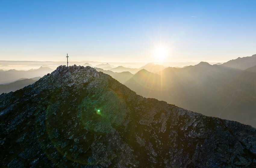 Rocky mountain ridge with summit cross at sunrise, sun over layered ranges and haze in the valleys | © Gasteiner Bergbahnen AG