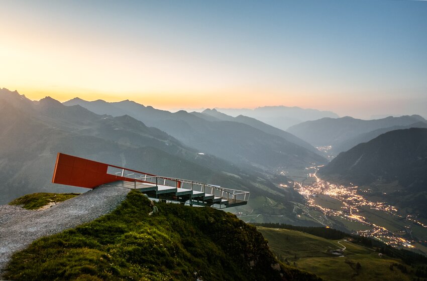 Modern viewing platform on mountain edge above illuminated valley with towns and winding road at dusk | © Gasteiner Bergbahnen AG