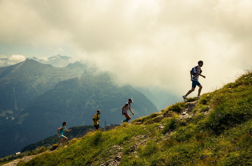 Vier Trailrunner laufen auf einem steilen Bergpfad durch alpine Wiesen vor hohen Bergen und Wolken. | © Michael Müller