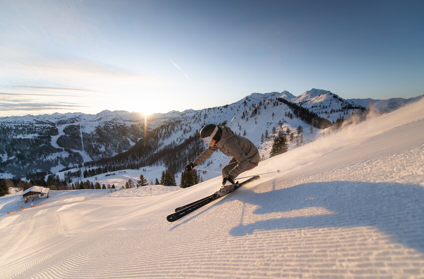 A skier carves a turn on a groomed slope in front of snow-covered mountains at sunrise under a clear sky. | © Christian Schartner
