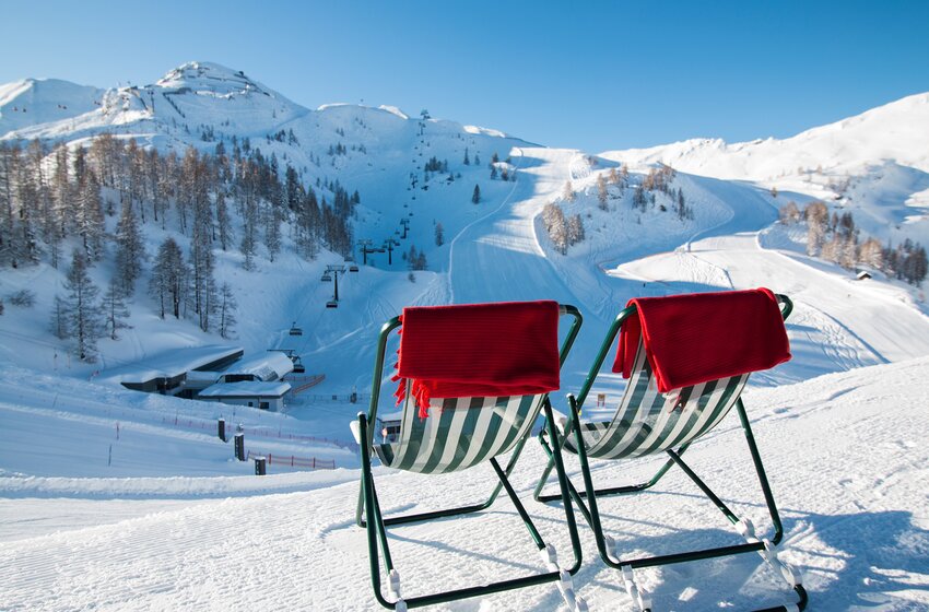 Two deck chairs with red blankets stand beside a snowy ski slope with lifts and snow-covered mountains. | © Zauchensee Liftgesellschaft Benedikt Scheffer GmbH