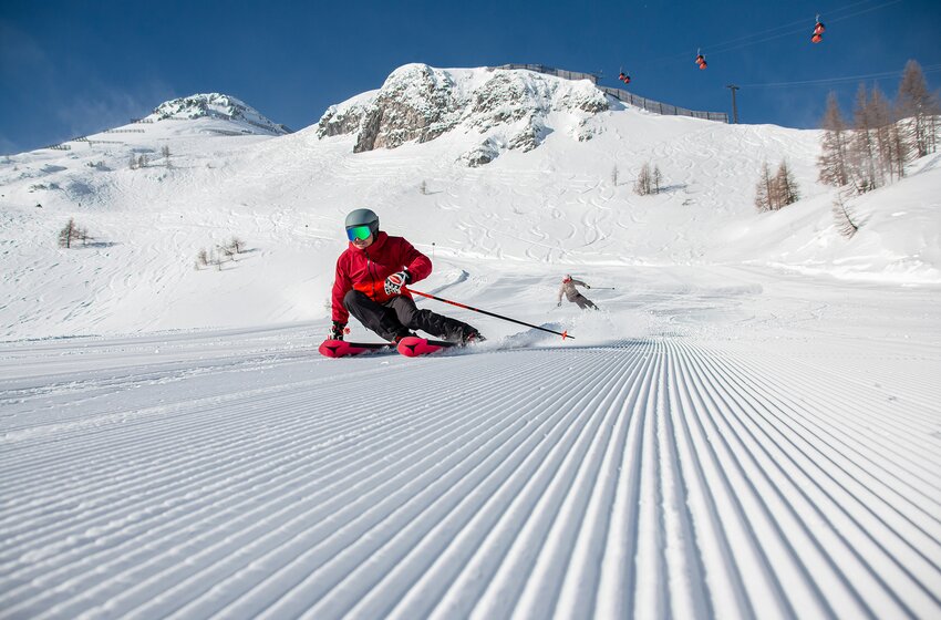 Zwei Skifahrer ziehen Schwünge auf einer präparierten Piste vor schneebedeckten Bergen und Gondelbahn bei Sonne. | © Zauchensee Liftgesellschaft Benedikt Scheffer GmbH