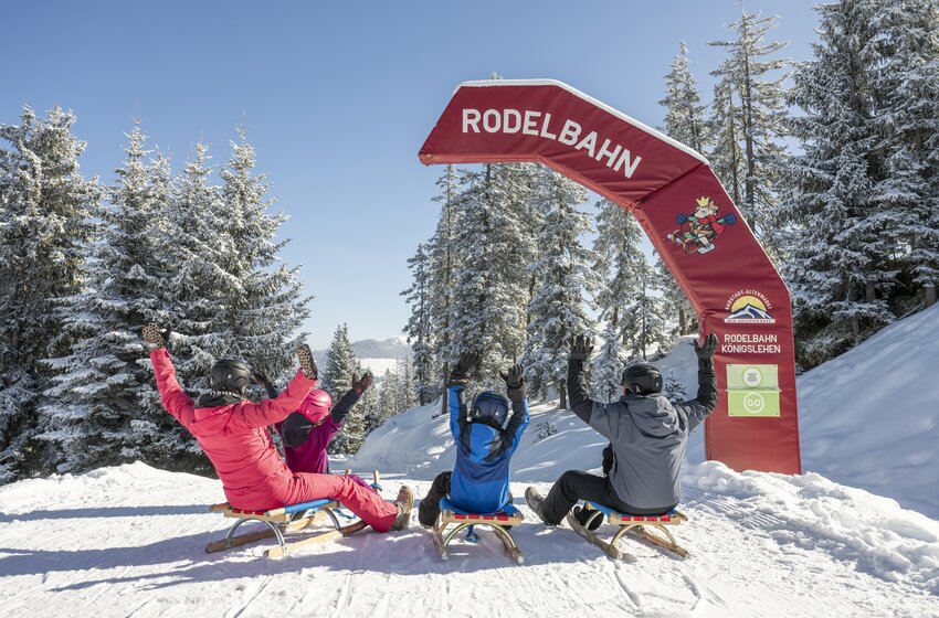 Drei Rodler fahren unter Startbogen der Rodelbahn Königslehen in verschneiten Winterwald mit erhobenen Armen | © Zauchensee Liftgesellschaft Benedikt Scheffer GmbH