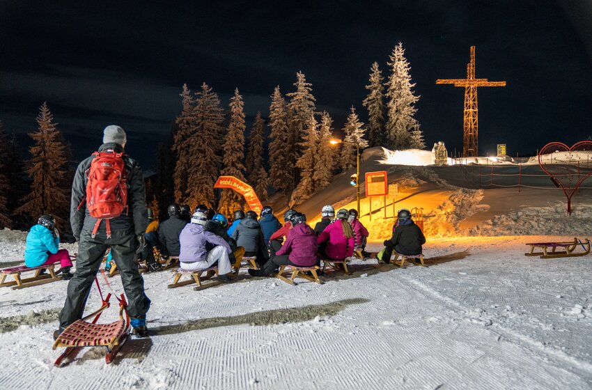 People on sleds sit at night in front of lit toboggan run with cross and snow-covered trees | © Zauchensee Liftgesellschaft Benedikt Scheffer GmbH